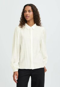 Blusa blanca de manga larga con botones en la parte delantera, cuello clásico y detalles fruncidos en los hombros; hecha de una tela ligera y texturizada.