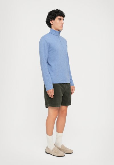 Camisa polo de manga longa azul clara com fecho de zíper, combinada com calções de algodão verde-escuro, meias brancas e mocassins bege.