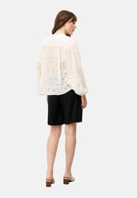 Doorzichtig off-white blouse met bloemenkantpatroon, lange pofmouwen en een ontspannen pasvorm, gecombineerd met zwarte shorts en lichtgekleurde blokhakken.