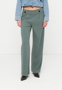 Weit geschnittene Hose aus gedämpft grünem Stoff mit glatter Textur, hoher Taille, vorderen Falten und zwei schwarzen Knopfakzenten.