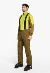 Skiausrüstung mit einem leuchtend grünen Langarmoberteil und braunen Hosen mit vorderen Trägern. Schwarze Skistiefel runden das Outfit ab.