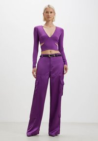 Crop top viola a maniche lunghe con scollo a V e dettagli cut-out, abbinato a pantaloni cargo a vita alta nella stessa tonalità, entrambi realizzati in tessuto morbido.
