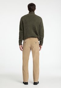 Ελιάς πράσινος, υφαντός σκούφος με ψηλό λαιμό, συνδυασμένος με ανοιχτό μπεζ παντελόνια από κοτλέ με πίσω τσέπες, φορεμένος με μαύρα παπούτσια.