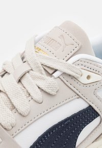 Světle béžové tenisky s bílými a námořnickými akcenty, texturovaný svršek z semiše a kůže, ploché tkané šňůrky a zlaté logo.