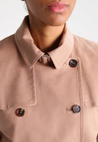 Jasnoróżowa kurtka z kołnierzykiem, charakteryzująca się podwójnym zapięciem, gładką fakturą, kontrastującymi guzikami i subtelnym połyskiem.