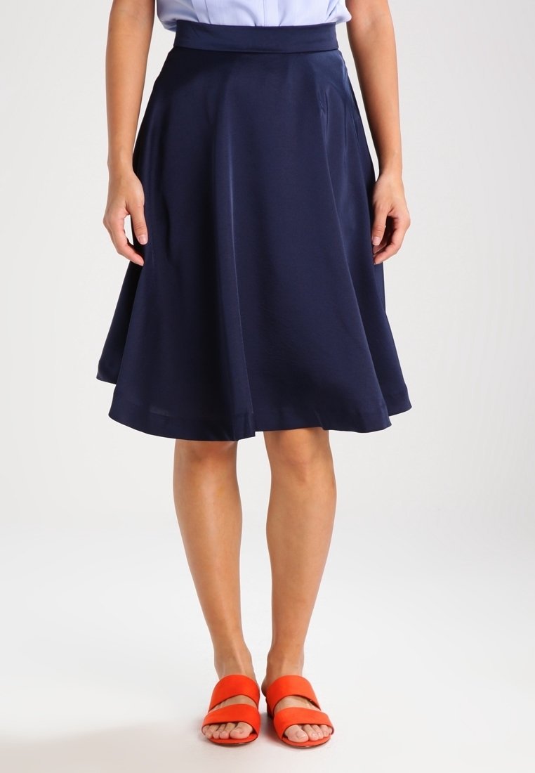 Femme portant une jupe bleu marine jusqu'aux genoux et des sandales ouvertes orange vif, debout avec les mains détendues le long du corps.