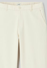 Pantalons couleur crème avec une texture de tissu lisse, une fermeture à bouton devant et des poches latérales, avec une coupe ajustée.