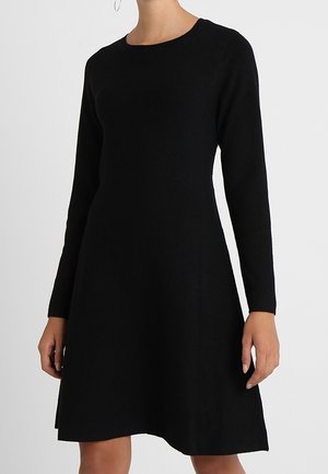 Femme portant une robe noire à manches longues, longueur genou, avec un col rond, debout sur un fond clair uni.
