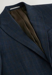 Blazer en tartan bleu marine avec un col à revers et deux poches avant. Doublure intérieure en marron avec des motifs géométriques. Texture douce, coupe ajustée.