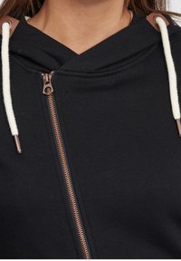 Felpa nera con zip e colletto alto, dotata di zip color rame e lacci crema, con dettagli in pelle color cuoio sulle spalle.
