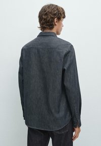 Dunkelblauses Jeanshemd mit langen Ärmeln, Kragen, geknöpften Manschetten, geradem Saum und strukturierter Oberfläche, von hinten gegen einen hellen Hintergrund gezeigt.
