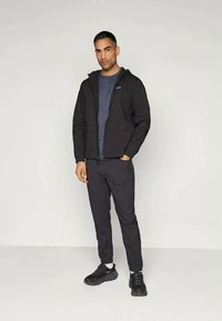Mand står mod en lys baggrund iført en sort zip-up hættetrøje, mørkegrå T-shirt, sorte bukser, hvide sokker og sorte sneakers.