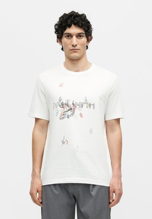 Νεαρός άνδρας με σγουρά σκούρα μαλλιά που φοράει λευκό μπλουζάκι με πολύχρωμο σχέδιο από διασκορπισμένους συνδετήρες και γκρι παντελόνι, στέκεται μπροστά σε απλό φόντο.