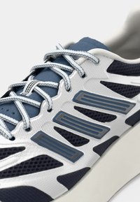 Sportssko med en hvid mesh-overdel, mørk navy detaljer, grå lag og lys blå snørebånd. Tekstureret sål for greb og stabilitet.
