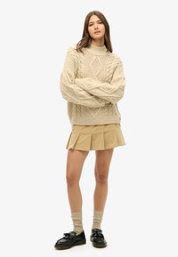 Beige strikket sweater med høj hals og kabelstrik mønster, parret med en tan plisseret nederdel. Sorte sko og lyse sokker fuldender looket.