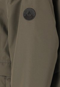 Olivgrön jackärm i slätt tyg. Har en cirkulär svart logotyp på ärmen med geometrisk design och text.