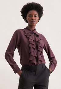 Langärmlige Bluse in dunklem Burgunderrot mit Rüschendetail an der Vorderseite, klassischem Kragen und glattem Stoff, kombiniert mit maßgeschneiderten schwarzen Hosen.