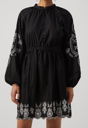 Czarna sukienka z długim rękawem, zapinana na guziki z przodu, z wiązaniem w pasie oraz białym kwiatowym haftem na rękawach i dole sukienki wykończonym falistym brzegiem.