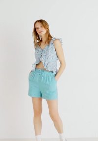 Světle modrý květinový crop top s volánkovými ramínky, doplněný tyrkysovými šortkami na stahování. Hladká tkanina s volným střihem, minimální kovové detaily.