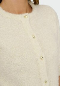 Gros pull en tricot de couleur blanc cassé doux avec un col rond et de petits boutons dorés brillants fixés à l'avant.