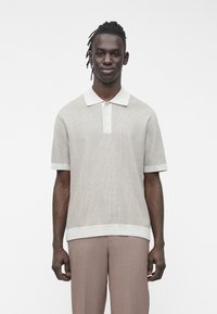 Muž s copánky má na sobě béžové texturované polo tričko s krátkým rukávem a světle hnědé kalhoty, stojící před jednoduchým bílým pozadím.