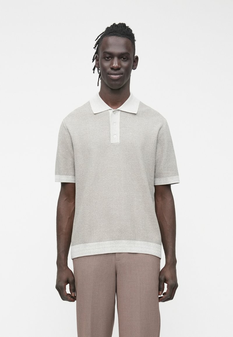 Muž s copánky má na sobě béžové texturované polo tričko s krátkým rukávem a světle hnědé kalhoty, stojící před jednoduchým bílým pozadím.