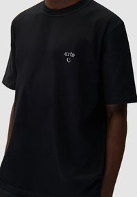 Maglietta nera in cotone con scollatura a girocollo, caratterizzata da un logo ricamato bianco e un grafico a forma di cuore sul lato sinistro del petto. Maniche corte.