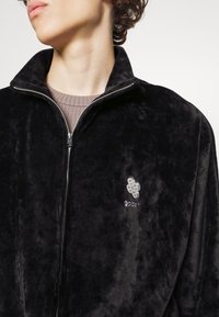 Zwarte, zachte jas met ritssluiting, hoge kraag en geborduurd logo. Zachte textuur, gestroomlijnd ontwerp, close fit en moderne stijlkenmerken.