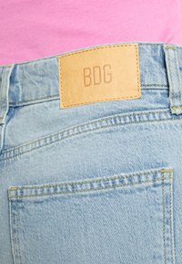 Os jeans de ganga de um azul claro apresentam um patch de couro com "BDG" em relevo. O tecido tem uma textura suave, com costuras visíveis e um bolso traseiro.