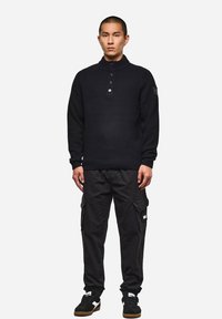 Schwarzer Strickpullover mit hohem Kragen und einer Dreiknopfknopfleiste, kombiniert mit Cargo-Hosen und schwarzen Sneakers. Mit gerippten Bündchen und Saum.