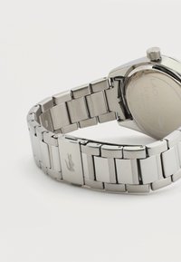 Ασημένιο ρολόι βραχιόλι με μεταλλικούς κρίκους και κλειστή αγκράφα, χαραγμένο με ένα μικρό λογότυπο κροκόδειλου σε λευκό φόντο.