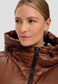 Braune gesteppte Jacke mit hohem Kragen und schwarzem Reißverschluss. Verfügt über Kordeln an der Kapuze und eine glatte, glänzende Textur.