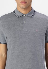 Īsām piedurknēm polo krekls, izgatavots no teksturēta pelēka auduma ar rakstainu apkakli, divām pogām un mazu logotipu uz krūtīm.