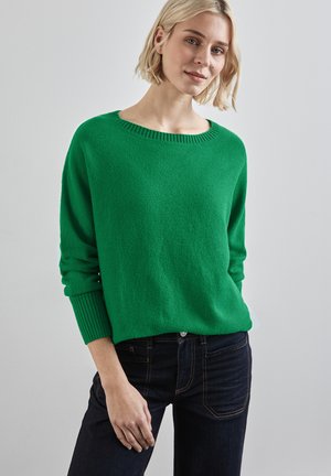Groene gebreide trui met een brede halslijn, geribbelde manchetten en een relaxte pasvorm, gecombineerd met donkere spijkerjeans. Gladde textuur en heldere kleur.