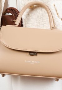 Sac à main en cuir beige avec un design structuré, une poignée supérieure et une fermeture à fermoir. Présente un logo métallique et des détails de couture.