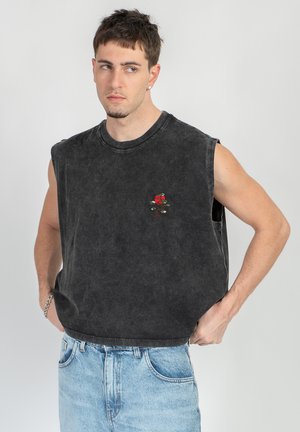 Giovane uomo con capelli corti che indossa una maglietta nera senza maniche con un ricamo di una rosa rossa e jeans azzurri, mani sui fianchi, che guarda di lato.