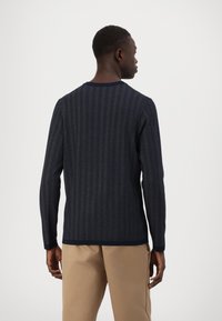 Maglione a maniche lunghe blu navy con motivo a spina di pesce; colletto e polsini a coste; texture morbida; vista posteriore; indossato con pantaloni beige.