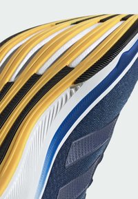 Gros plan d’une semelle de chaussure de sport bleue avec une semelle intermédiaire blanche, un liseré bleu, des bandes de traction en caoutchouc noires et jaunes, et une tige en tissu texturé.