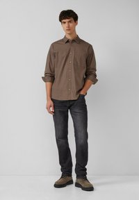 Junger Mann, der ein braun gemustertes Langarmhemd, dunkle Jeans und braune Schnürschuhe trägt, vor einem schlichten, hellen Hintergrund steht.