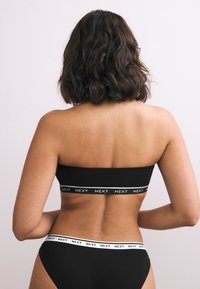 Frau von hinten gesehen, trägt schwarzen Bandeau-Top und Slips mit weißen Bändern mit der Aufschrift "NEXT" auf einfachem hellem Hintergrund.