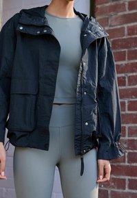 Schwarze leichte Jacke mit Kapuze, Reißverschluss und Druckknöpfen, mit elastischen Bündchen und einer strukturierten Oberfläche, kombiniert mit einem eng anliegenden grauen Oberteil und Leggings.