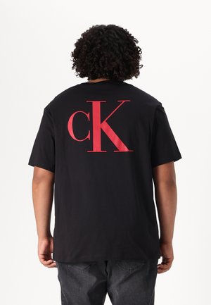 Person mit lockigem Haar trägt ein schwarzes T-Shirt mit großem roten "cK"-Logo auf dem Rücken und dunkelgraue Hose vor weißem Hintergrund.