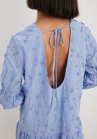 Light blue, long-sleeved dress with floral embroidery, open back, and tie closure at neckline; textured fabric with puffed sleeves.
