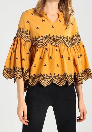 Hořčicově žlutá halenka s rozšířenými rukávy, černými vyšívanými květinovými a hvězdnými vzory, vroubkovaným lemem a lehkou tkaninou.