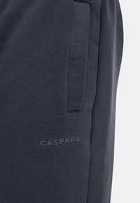 Mørkegrå joggebukser laget av myk stoff, med en sidelomme og preget "CASPARA"-logo nær den nederste sømmen.