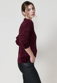 Burgunderfarbener Strickpullover mit strukturiertem Muster, gerippten Bündchen und Saum. Kombiniert mit schwarzen, eng anliegenden Hosen. Seitenansicht, neutraler Hintergrund.