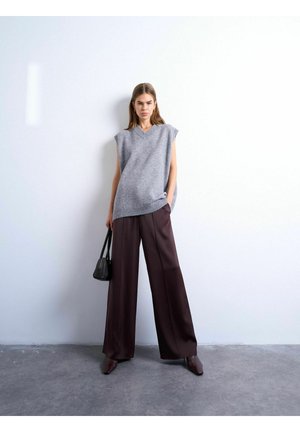 Femme portant un pull sans manches en maille grise, un pantalon large marron et des bottines, debout contre un mur blanc tenant un sac à main noir.