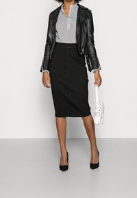 Zwarte aansluitende kokerrok gecombineerd met een wit-zwart gebloemde blouse en een zwarte leren motorjas. Houdt een witte handtas vast.