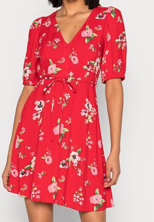Frau trägt ein rotes Blumenkleid mit kurzen Puffärmeln, V-Ausschnitt, Knopfdetails und gebundenem Taillengürtel.