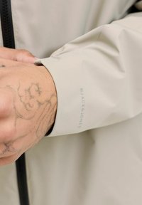 Veste imperméable beige clair avec une texture lisse, des poignets réglables et un logo visible sur la manche. Fermeture avant à glissière.